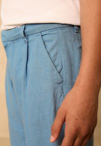 Pantalon en lin bleu clair présenté, avec plis avant, poches latérales et finition texturée. La main du modèle est visible sur le côté.
