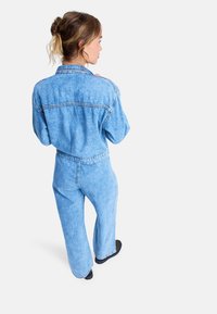 Mono de mezclilla azul claro con un ajuste holgado, perneras anchas, mangas largas y un diseño de costura expuesta, que cuenta con una cintura ceñida por un cinturón y un cuello.