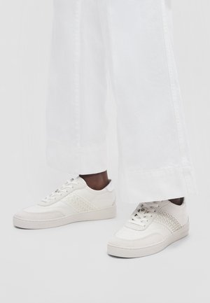 Adidași albi cu design cu șireturi și panouri laterale cu model, purtați cu pantaloni albi largi.