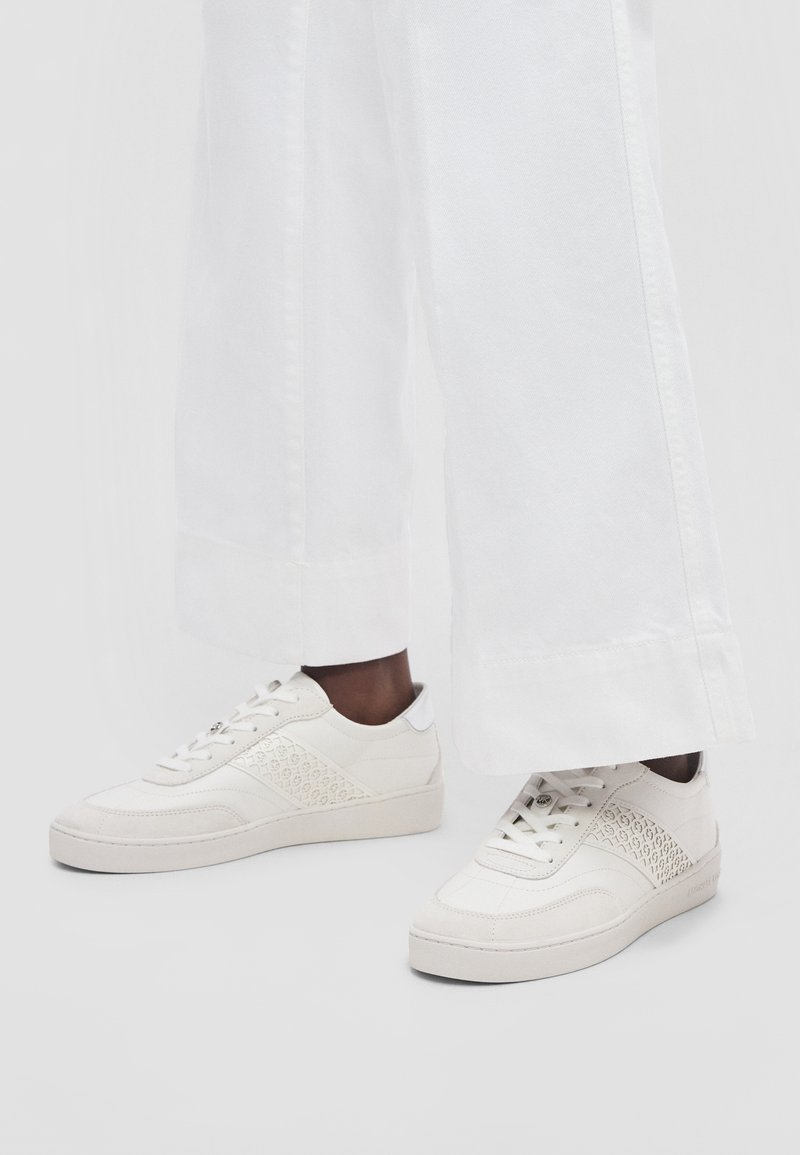 Baskets blanches avec un design à lacets et des panneaux latéraux à motifs, portées avec un pantalon blanc à jambes larges.