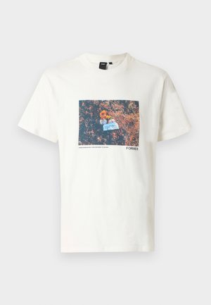 T-shirt en coton crème avec une impression graphique rectangulaire de fleurs et d'un objet bleu sur un fond brun texturé. Étiquette noire au niveau du col.