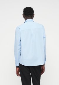 Ανοιχτό μπλε πουκάμισο μακρυμάνικο με γιακά με κουμπιά, λεπτομέρεια πλάτης με πιέτα και κουμπιά στα μανίκια. Λείο βαμβακερό ύφασμα, εφαρμοστή γραμμή.