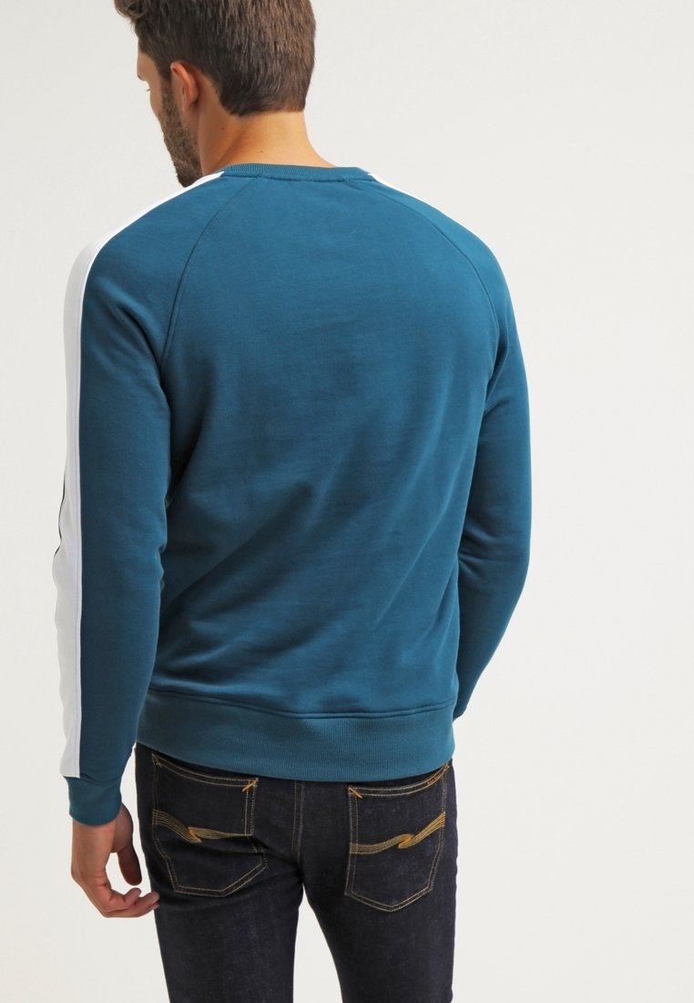 Homme portant un sweat-shirt bleu sarcelle avec des bandes blanches sur les manches et un jean foncé, vu de dos sur un fond uni.