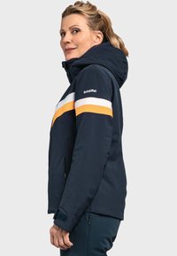 Marineblaue isolierte Jacke mit Kapuze, ausgestattet mit orangefarbenen und weißen Streifen an den Schultern. Das Material wirkt glatt und hat Reißverschlusstaschen.