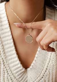 Okrągły metalowy naszyjnik z wisiorem o geometrycznym wycięciu, zawieszony na delikatnym łańcuszku. Ręka modelki wskazuje na wisiorek.