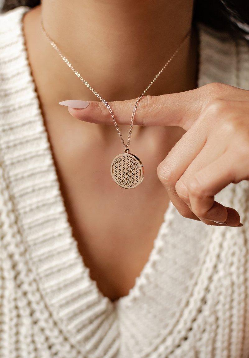 Okrągły metalowy naszyjnik z wisiorem o geometrycznym wycięciu, zawieszony na delikatnym łańcuszku. Ręka modelki wskazuje na wisiorek.