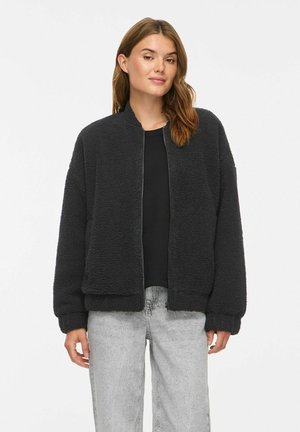 Femme aux cheveux châtain clair portant une veste polaire noire zippée, un t-shirt noir et un jean gris clair, debout devant un fond blanc uni.