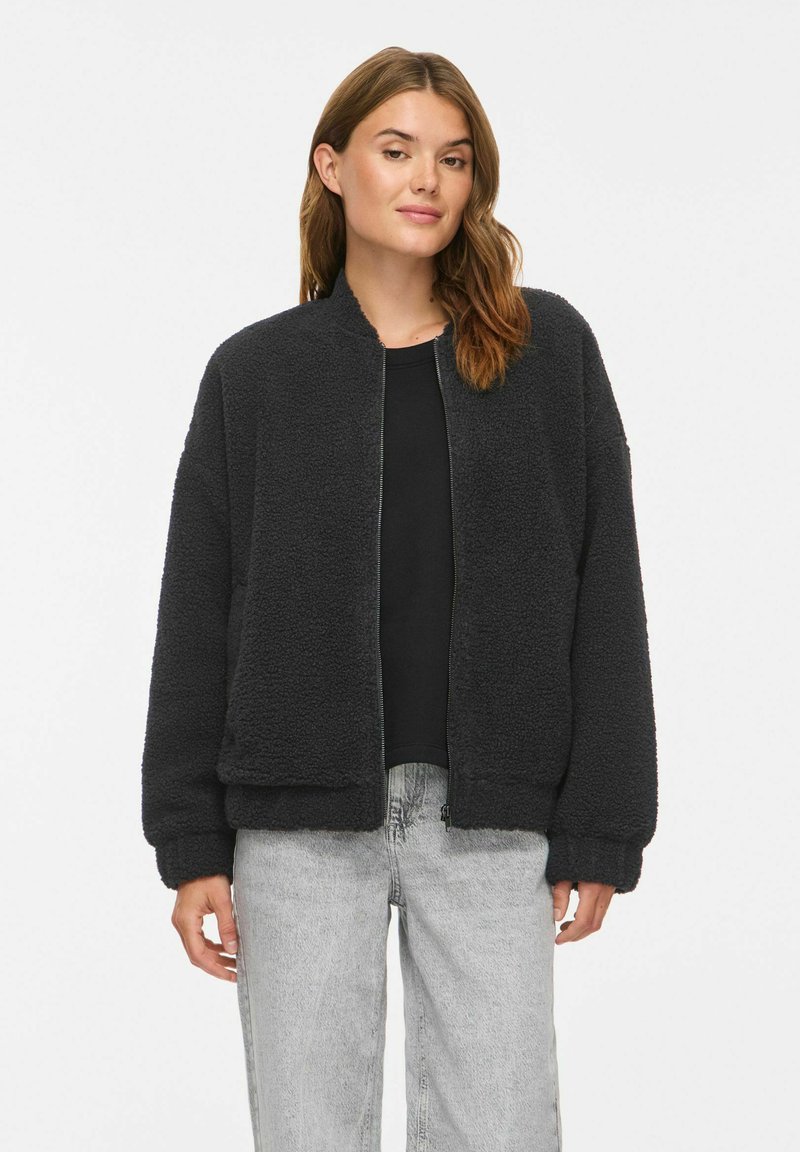 Femme aux cheveux châtain clair portant une veste polaire noire zippée, un t-shirt noir et un jean gris clair, debout devant un fond blanc uni.