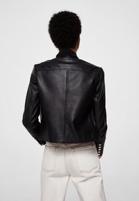 Musta nahkatakki, jossa on istuva malli, hienovarainen kiilto, lyhyt pituus ja napit hihansuissa. Valkoiset housut näkyvissä.