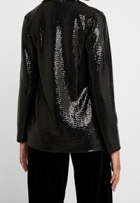 Černý blazer s flitry, lesklou texturou a strukturovaným designem. Dlouhé rukávy, klopový límec a volný střih, doplněný černými kalhotami.