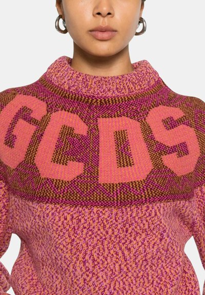 Donna che indossa un maglione lavorato a maglia rosa e marrone con le lettere "GCDS" in grandi lettere a blocchi sul petto.