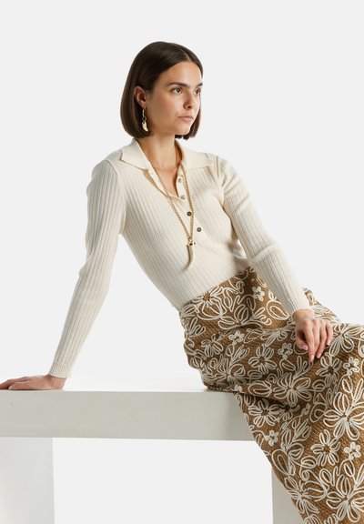 Cremfarbener, gerippter Langarm-Top mit einem Polo-Kragen und Knopfdetails, kombiniert mit einem braunen Rock mit floraler Stickerei und weißen Akzenten.
