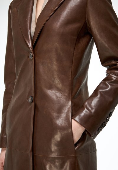 Nő, barna, alakra szabott bőrblézerben, két gombbal, hajtókával, az egyik kezét a zakózsebbe dugva.