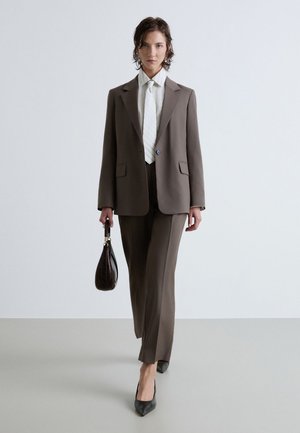 Femme marchant vers l'avant portant un costume marron avec une chemise blanche et une cravate, tenant un sac à main foncé et portant des talons aiguilles noirs pointus sur un sol gris.