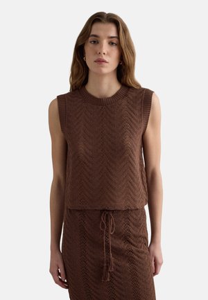 Donna che indossa un top marrone lavorato a maglia senza maniche e una gonna coordinata con coulisse, in piedi davanti a uno sfondo chiaro e uniforme.