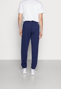 Person som bär marinblå Adidas sweatpants, vit t-shirt, vita strumpor och vita sneakers, ståendes vänd mot en enkel vägg.