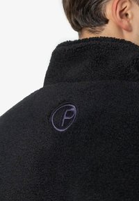 Mężczyzna w czarnej polarowej kurtce o strukturalnej fakturze, z podniesionym kołnierzem i haftowanym okrągłym logo na górnej części pleców.