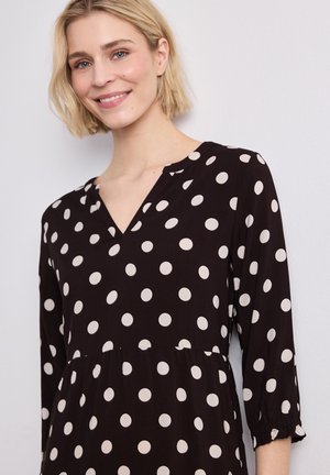 Femme blonde souriante, portant une robe noire à gros pois blancs et manches trois-quarts, debout contre un mur clair uni.