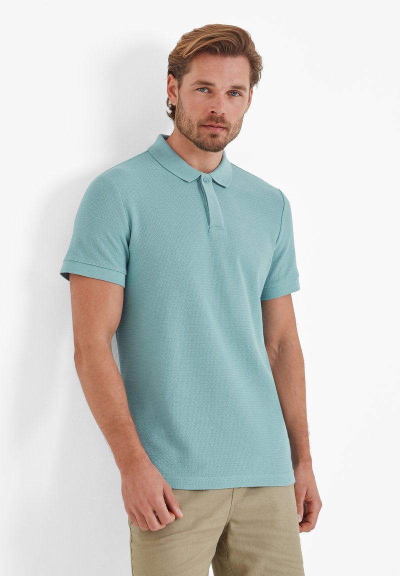 Polo shirt in light blue, made from textured fabric. Features a collar, short sleeves, and a two-button placket.