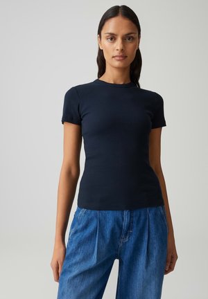 Donna con lunghi capelli scuri indossa una maglietta nera aderente a maniche corte e jeans di denim blu larghi, in piedi contro uno sfondo semplice e chiaro.