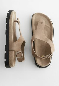 Bruine suède sandalen hebben een teengeleide ontwerp met een verstelbare band en een gestructureerde zwarte rubberen zool, die comfort en grip benadrukken.