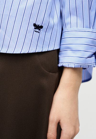 Κοντινή λήψη του μανικιού και του τελειώματος ενός μπλε και λευκού ριγωτού πουκάμισου με μαύρο κέντημα πεταλούδας, φορεμένο με σκούρο καφέ παντελόνι και ένα χέρι σε ηρεμία.