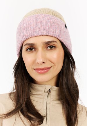 Femme aux longs cheveux bruns portant une veste beige et un bonnet en tricot rose et beige, souriant doucement sur fond blanc.