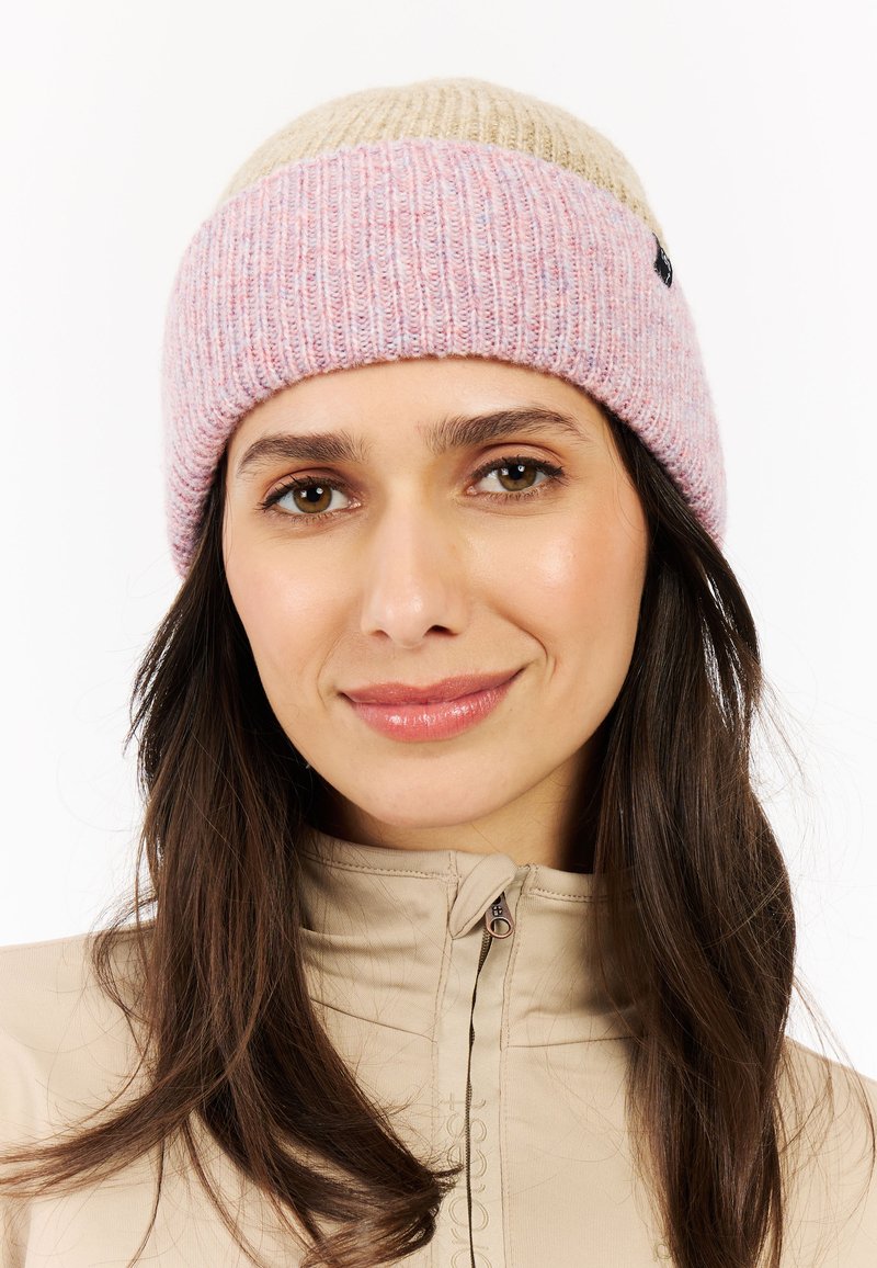 Vrouw met lang bruin haar, gekleed in een beige jasje en een roze en beige gebreide muts, die vriendelijk glimlacht tegen een witte achtergrond.