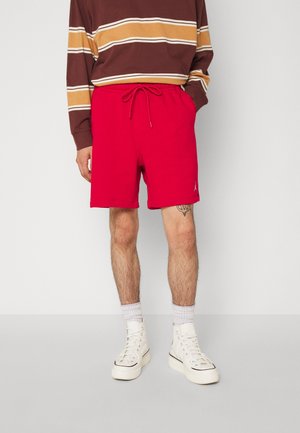 Persona que lleva pantalones cortos rojos con cordón, zapatillas blancas de caña alta, calcetines blancos y una camiseta de manga larga marrón a rayas, de pie frente a un fondo liso.