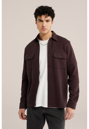 Junger Mann mit lockigem Haar, der ein dunkelbraunes Hemd mit Knöpfen über einem weißen T-Shirt und dunklen Jeans vor einem hellbeigen Hintergrund trägt.