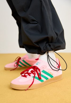 Osoba w czarnych spodniach z regulowanymi mankietami i białych skarpetkach, różowe buty sportowe Adidas Gazelle z zielonymi paskami i czerwonymi sznurówkami na żółtej podłodze.