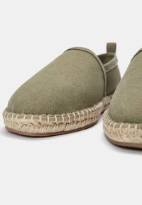 Chaussures slip-on en toile verte avec un détail en corde de jute autour de la semelle et une base plate en caoutchouc, affichant un design minimaliste et des accents de couture.