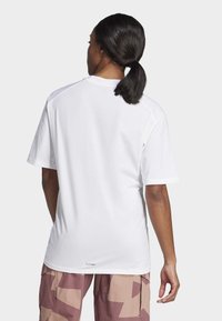 Bílý tričko s krátkým rukávem a volným střihem, hladkou texturou a diskrétní logem blízko spodního lemu, v kombinaci s vzorovanými, světle hnědými cargo kalhotami.