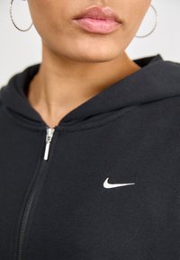 Närbild av en kvinnas nedre ansikte som bär silverfärgade hoopörhängen och en svart Nike zip-up hoodie med en vit swoosh-logotyp.