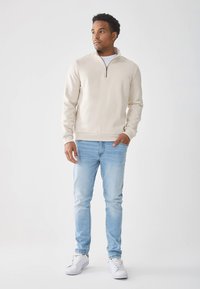 Béžová mikina na zips s dlhými rukávmi, nosená cez bielu košeľu, skombinovaná so svetlomodrými džínsami a bielymi teniskami. Minimalistický dizajn.