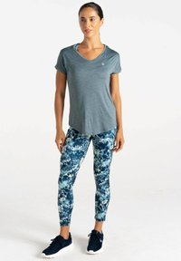 Camiseta atlética gris con cuello en V; leggings azules con un diseño abstracto; modelo vistiendo zapatillas negras, de pie en un fondo neutro.