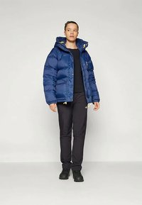 Ženska v modri podloženi jakni z rumenimi zaponkami, črnih hlačah in črnih čevljih, stoji pred enostavno svetlo ozadje.