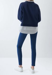 Felpa blu scuro sovrapposta a una maglietta a righe, abbinata a jeans skinny blu e sneakers bianche con un accento grigio sulla scarpa.