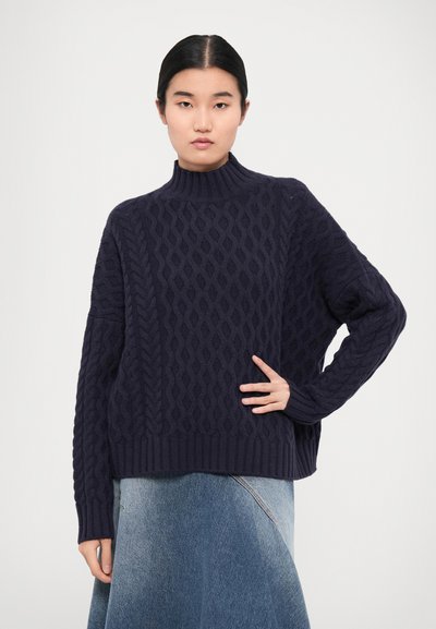 Dark navy cable-knit sweater with a high neckline, featuring textured pattern and ribbed cuffs. Paired with a denim skirt.
