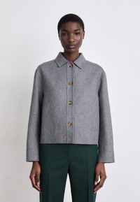 Casaco de lã cinza com colarinho, equipado com cinco botões redondos dourados e mangas de corte reto; complementado por calças verde de corte elegante.