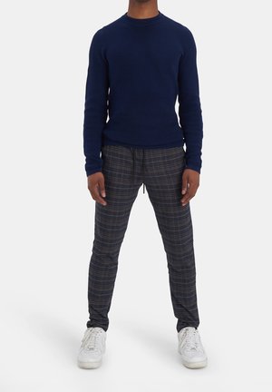 Persona che indossa un maglione blu navy, pantaloni grigi a quadri con coulisse e sneakers bianche, in piedi davanti a uno sfondo semplice.