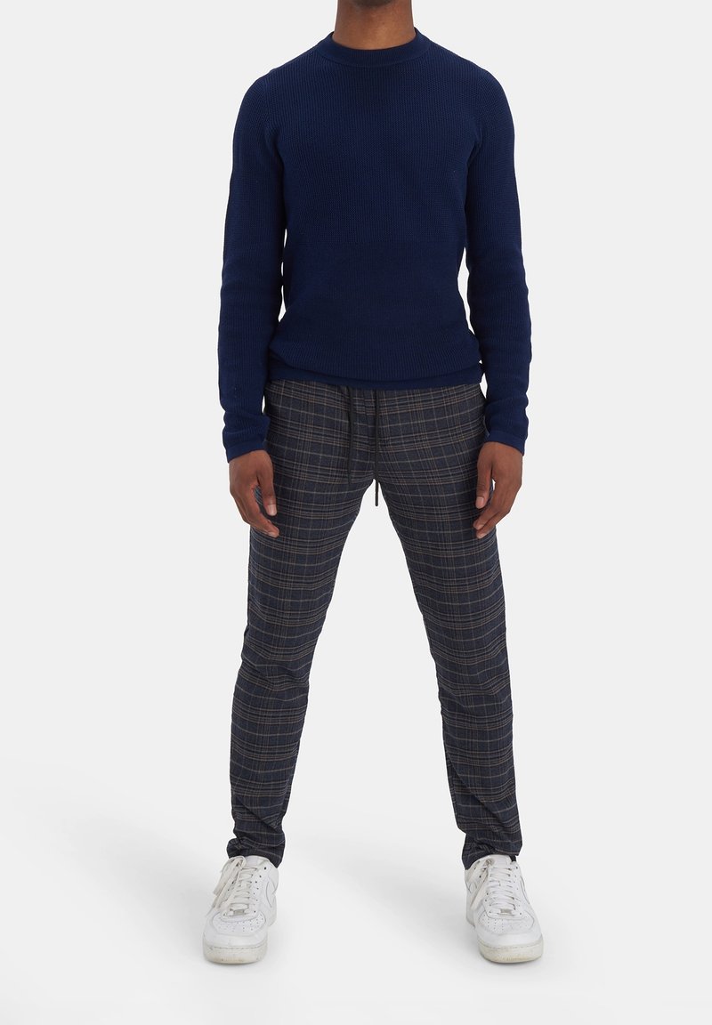 Personne portant un pull bleu marine, un pantalon gris à carreaux avec cordon et des baskets blanches, debout devant un fond uni.
