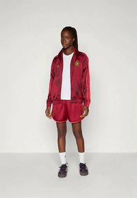 Bordeauxfarbene Sporttrainingsanzug mit roten Akzenten. Satin-Jacke mit Streifen, besticktem Emblem. Weißes T-Shirt, rote Shorts, weiße Socken und schwarze Schuhe.