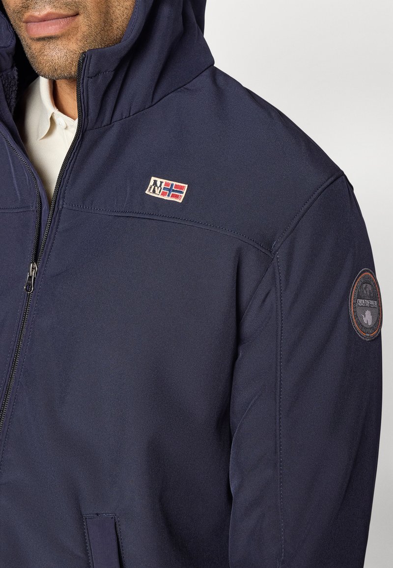 Chaqueta azul marino con capucha, que presenta un parche de logo en el pecho y un emblema circular en la manga. Textura suave y cierre con cremallera.