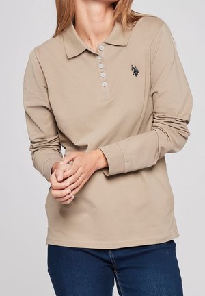 Kobieta ubrana w beżową koszulkę polo z długim rękawem z guzikami i ciemnym wyszywanym logo, w połączeniu z niebieskimi dżinsami, ręce splecione z przodu.