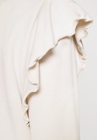 Top beige claro con una textura suave, que presenta mangas con volantes y un diseño simple. La tela parece lisa y ligeramente elástica.