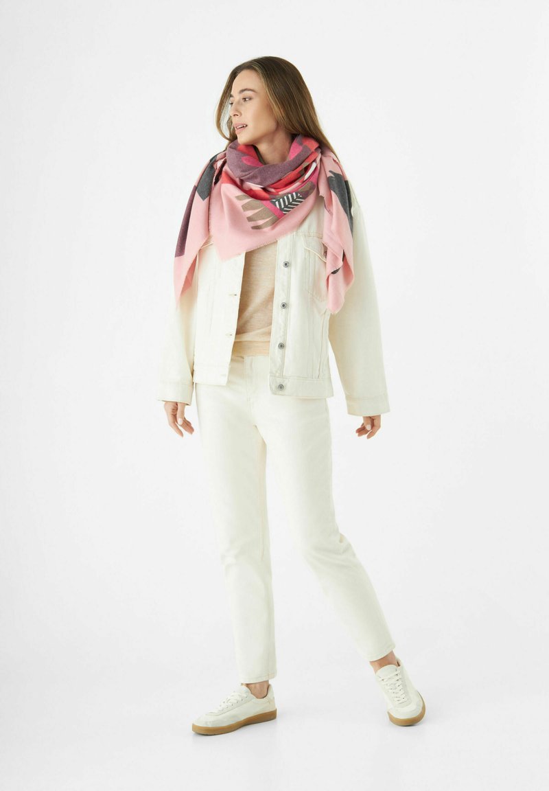 Écharpe plaid en rose, gris et noir avec des motifs géométriques, portée sur un haut beige clair et une veste en jean blanche, ainsi que des pantalons blancs.
