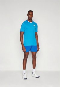 Blå atletisk t-skjorte med kontrasterende hvite kanter, kombinert med blå shorts. Materialet virker lett. Modellen har på seg hvite joggesko.