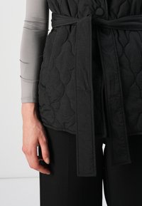 Gilet noir matelassé avec une taille nouée, doté d'une fermeture à boutons et d'un motif texturé. Porté par-dessus un haut gris à manches longues.