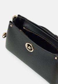 Bolso de piel texturizada negra tipo crossbody con herrajes en tono dorado, cierre de cremallera y un distintivo logo circular en la parte frontal.